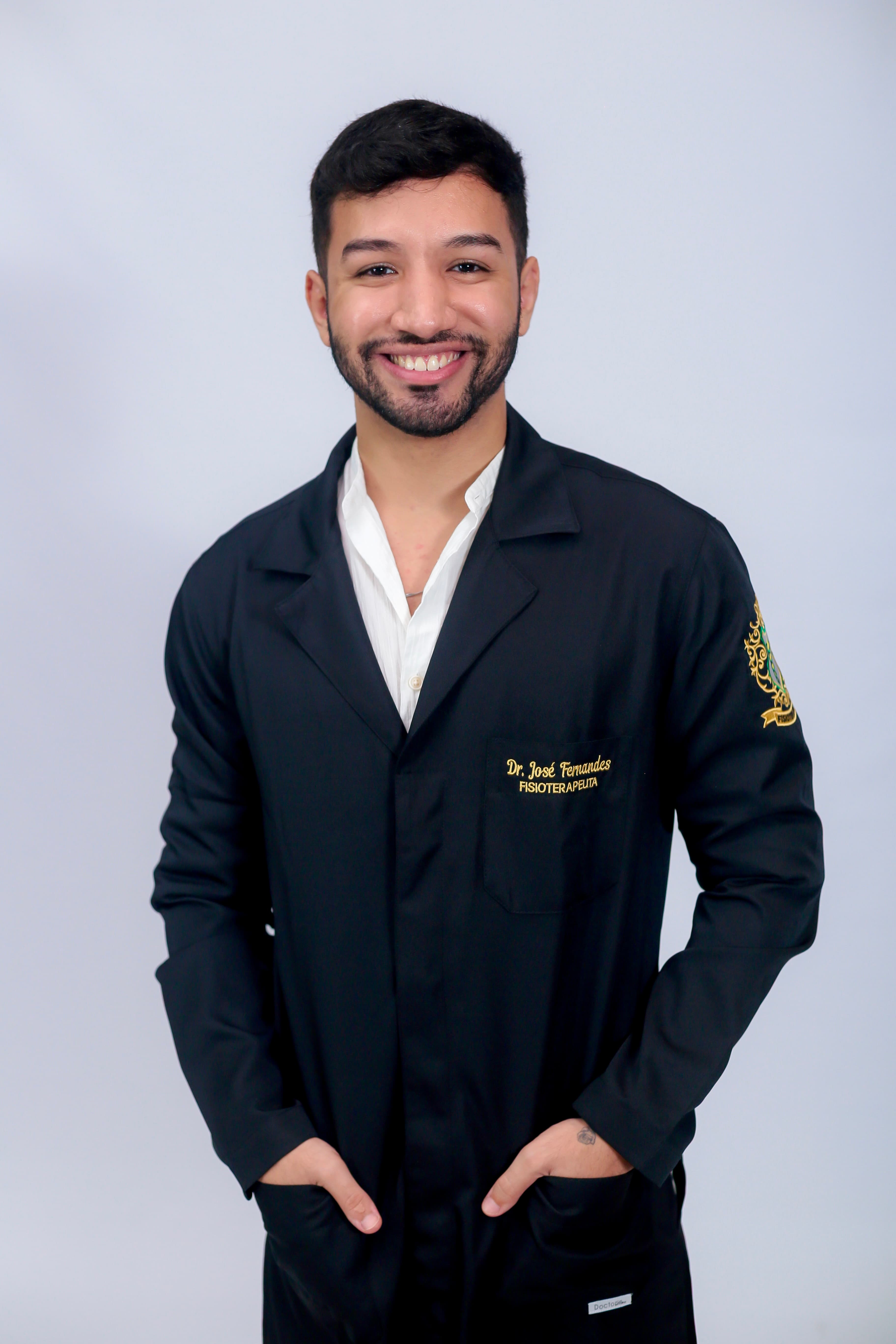 Dr. José Fernandes sorrindo, de jaleco preto com o nome e a palavra Fisioterapeuta bordados em dourado no bolso